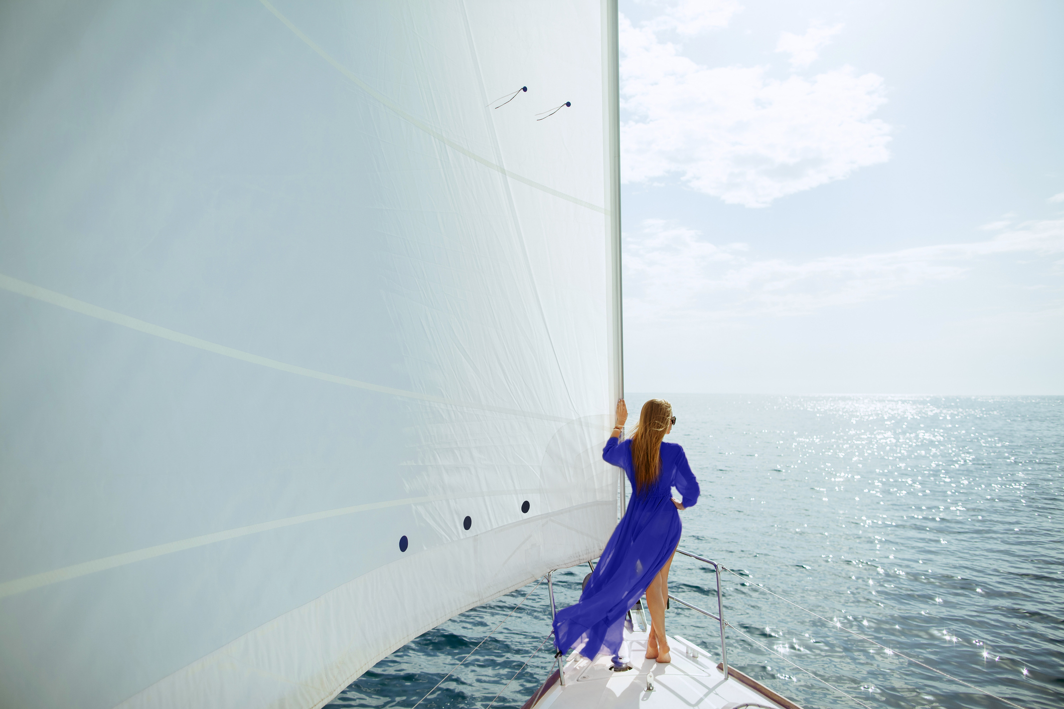 Femme en robe bleue de dos sur un voilier, lumière d'été et mer scintillante : registre visuel d'une campagne d'été.